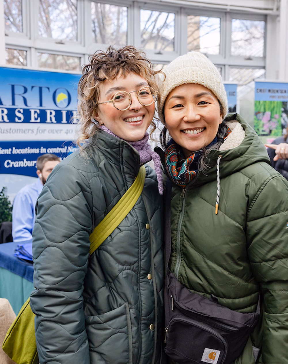 Two women smiling together in a brightly lit indoor setting. Both wear warm jackets; one has curly hair and glasses, and the other has straight hair under a cream-colored beanie. A display banner is visible in the background.