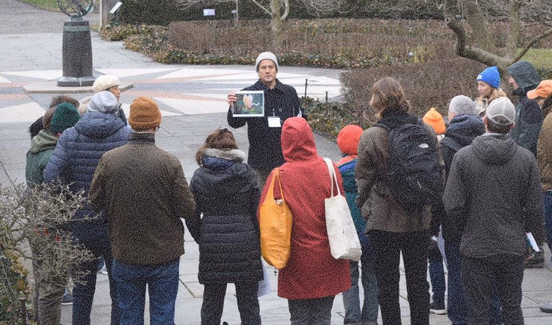A tour guide holding a photograph stands in front of a group of people who are gathered around her in a park. The attendees are dressed in winter clothing, and the landscape features trees and a statue in the background.