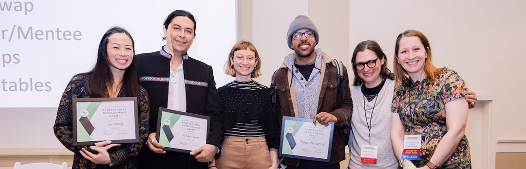 A diverse group of five individuals stands together, each holding certificates. The people include two women and three men, dressed in semi-formal attire, smiling. A presentation backdrop is visible with text that includes 'Mentor/Mentee'.