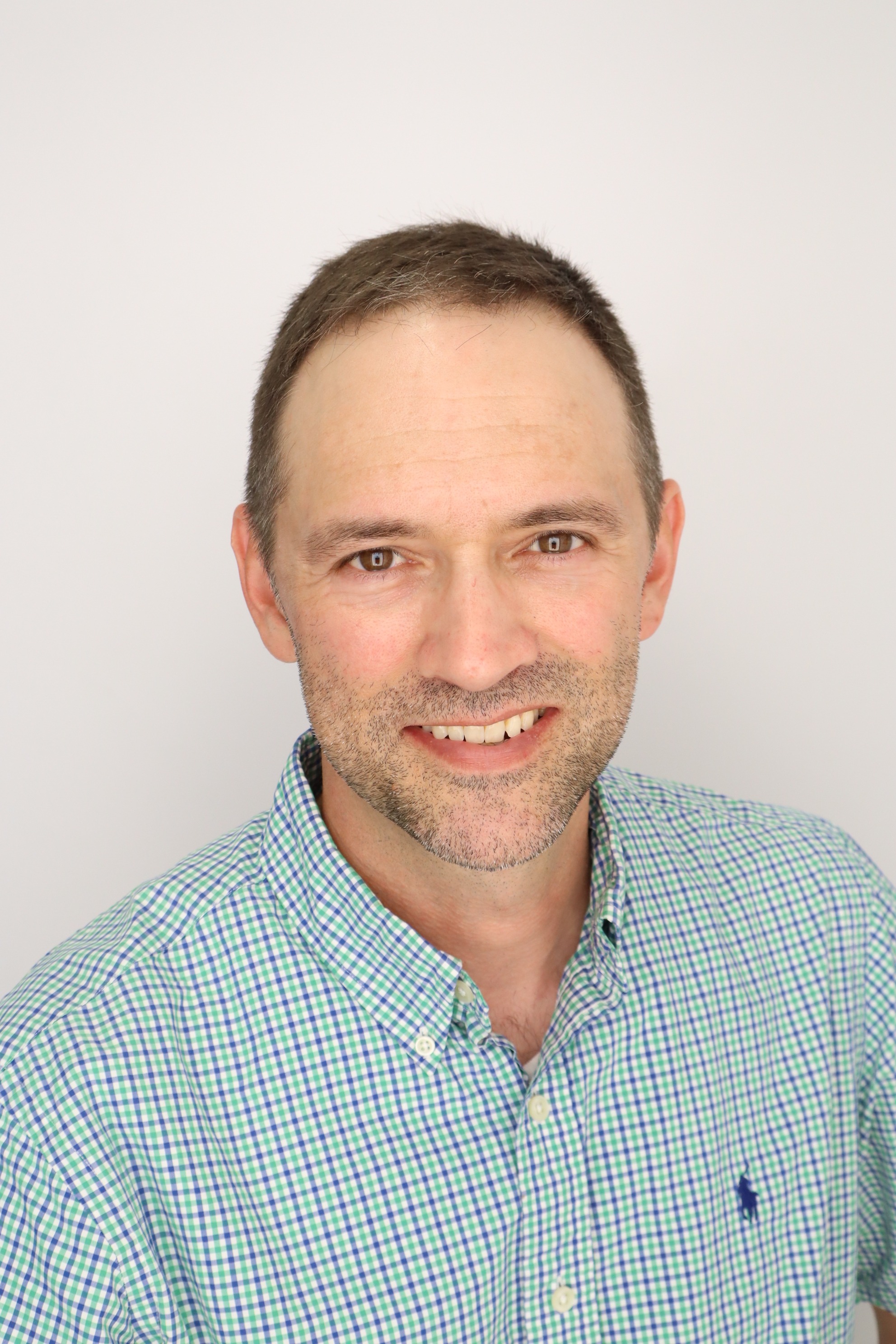 A man with short brown hair and a light beard smiles at the camera. He is wearing a green and white checkered shirt. The background is plain white, enhancing the focus on the subject.