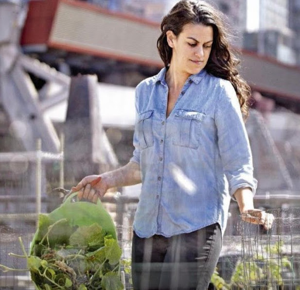A woman with long dark hair wearing a light blue button-up shirt stands in a garden bed, holding a green bag filled with plants. She looks down while tending to the garden, with buildings in the background.